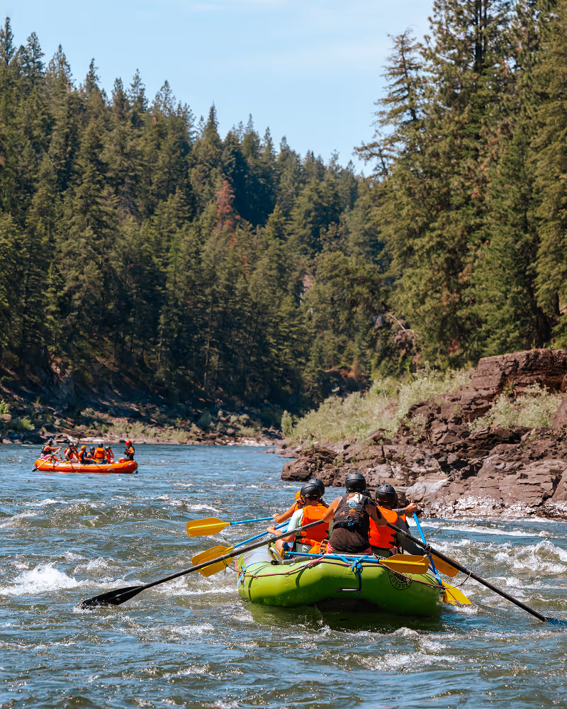 People wearing helmets and life vests rafting on a river surrounded by evergreen trees and rocky banks under clear sky.