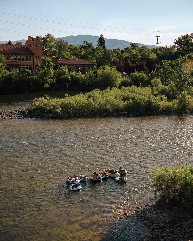 Group of people floating on inner tubes on a calm river surrounded by trees and buildings with mountains in the background.