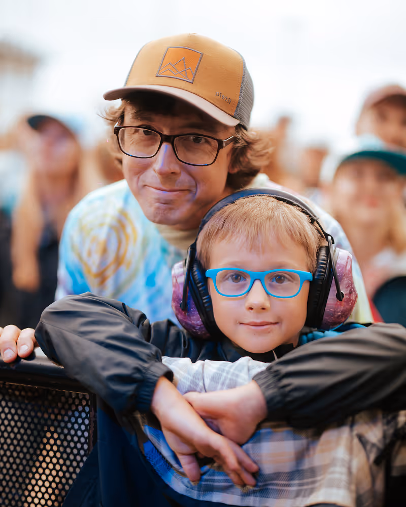 Man in a brown cap and glasses smiling behind a boy with blue glasses and noise-canceling headphones at an outdoor event.
