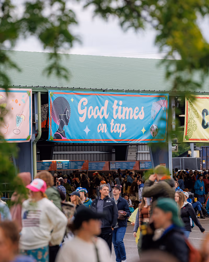 Crowd of people at an outdoor event beneath a blue banner reading 'Good times on tap' with artistic illustrations.