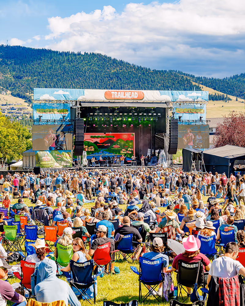 Outdoor music festival with a large crowd seated on colorful chairs facing a stage labeled 'Trailhead' against a backdrop of forested mountains and blue sky.