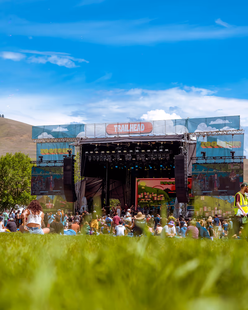 Outdoor music festival stage labeled 'Trailhead' with a crowd seated on grass under blue sky.