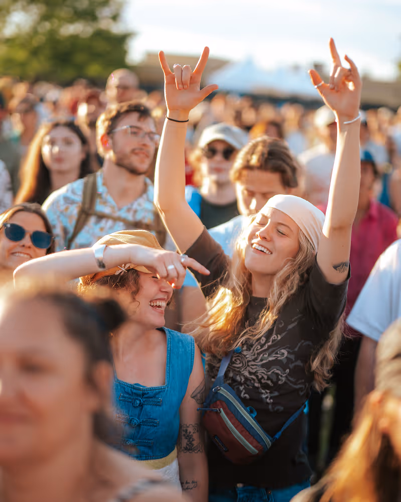 Two young women enjoying an outdoor concert, one smiling and wearing a bandana with raised hands making rock signs.