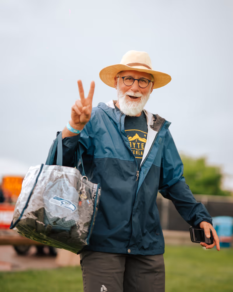 Smiling elderly man with white beard and glasses wearing a hat and blue jacket, holding a silver Seahawks bag and making a peace sign.