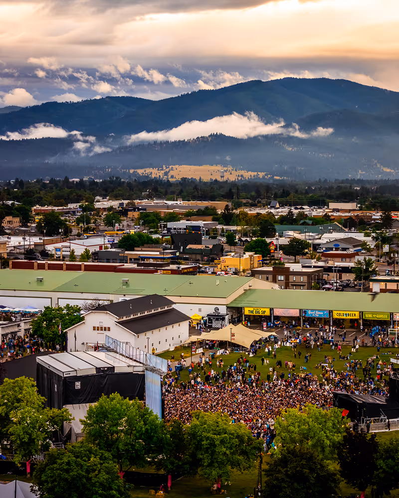Large outdoor concert crowd gathered in front of a stage with mountain range and cloudy sky in the background.