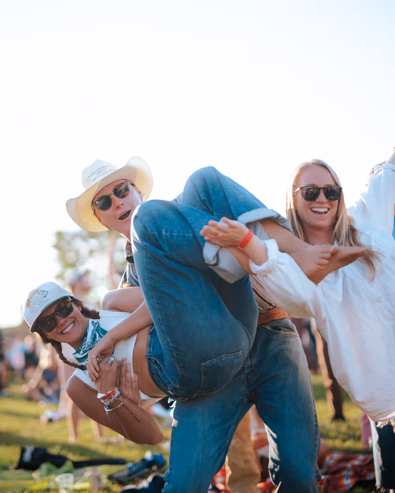 Three friends outdoors at a festival, one person wearing a white cowboy hat and sunglasses lifting another smiling person in sunglasses and a cap, with a third person in sunglasses and a white shirt cheering.