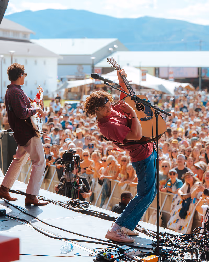 Two musicians playing guitar on an outdoor stage in front of a large, enthusiastic crowd during daytime.