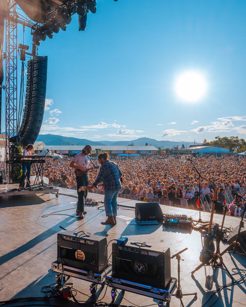 Outdoor concert stage with musicians playing guitars and keyboard as a large crowd watches under a bright sun and clear blue sky.