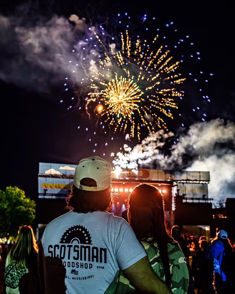 Couple watching colorful fireworks at night with a crowd and stage in the background.