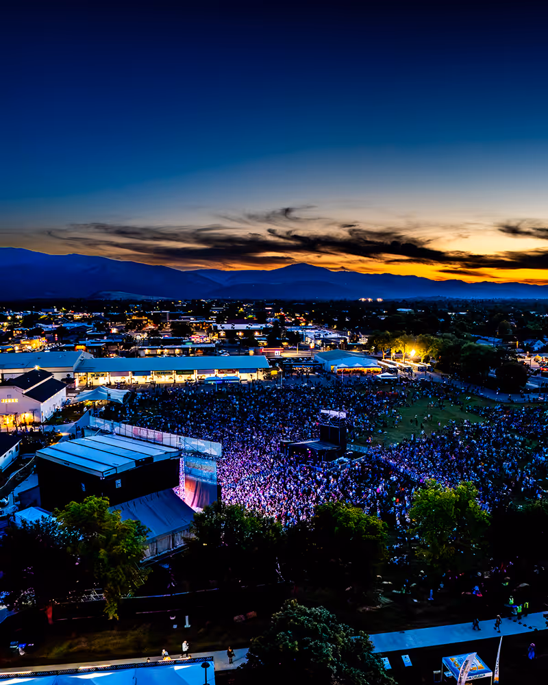 Aerial view of a large outdoor concert with a crowd in front of a stage at dusk, mountains and a colorful sunset in the background.