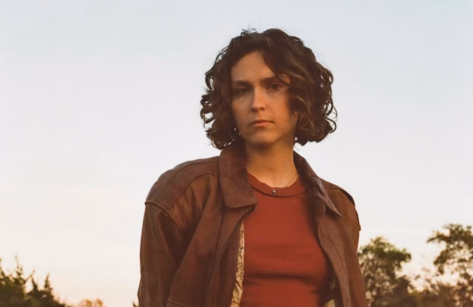 Young woman with curly brown hair wearing a brown jacket and red shirt outdoors at sunset.