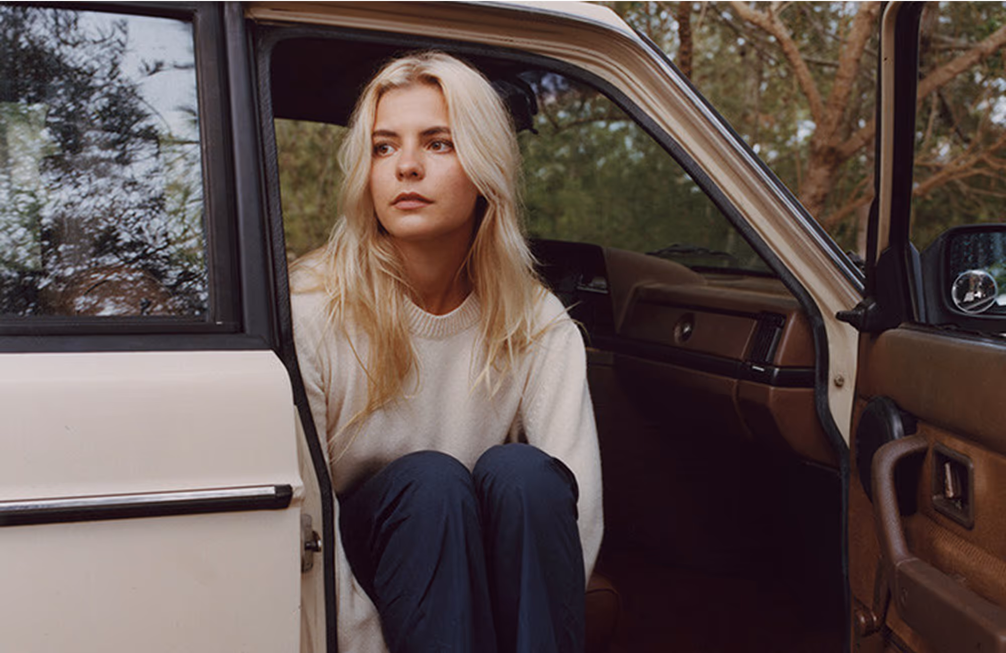 Young woman with long blonde hair sitting in the open doorway of a vintage car, looking contemplative.