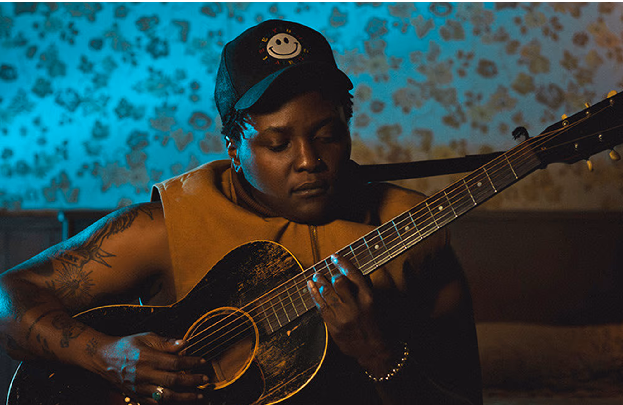 Person wearing a sleeveless jacket and cap playing an acoustic guitar indoors with a blue patterned wall in the background.