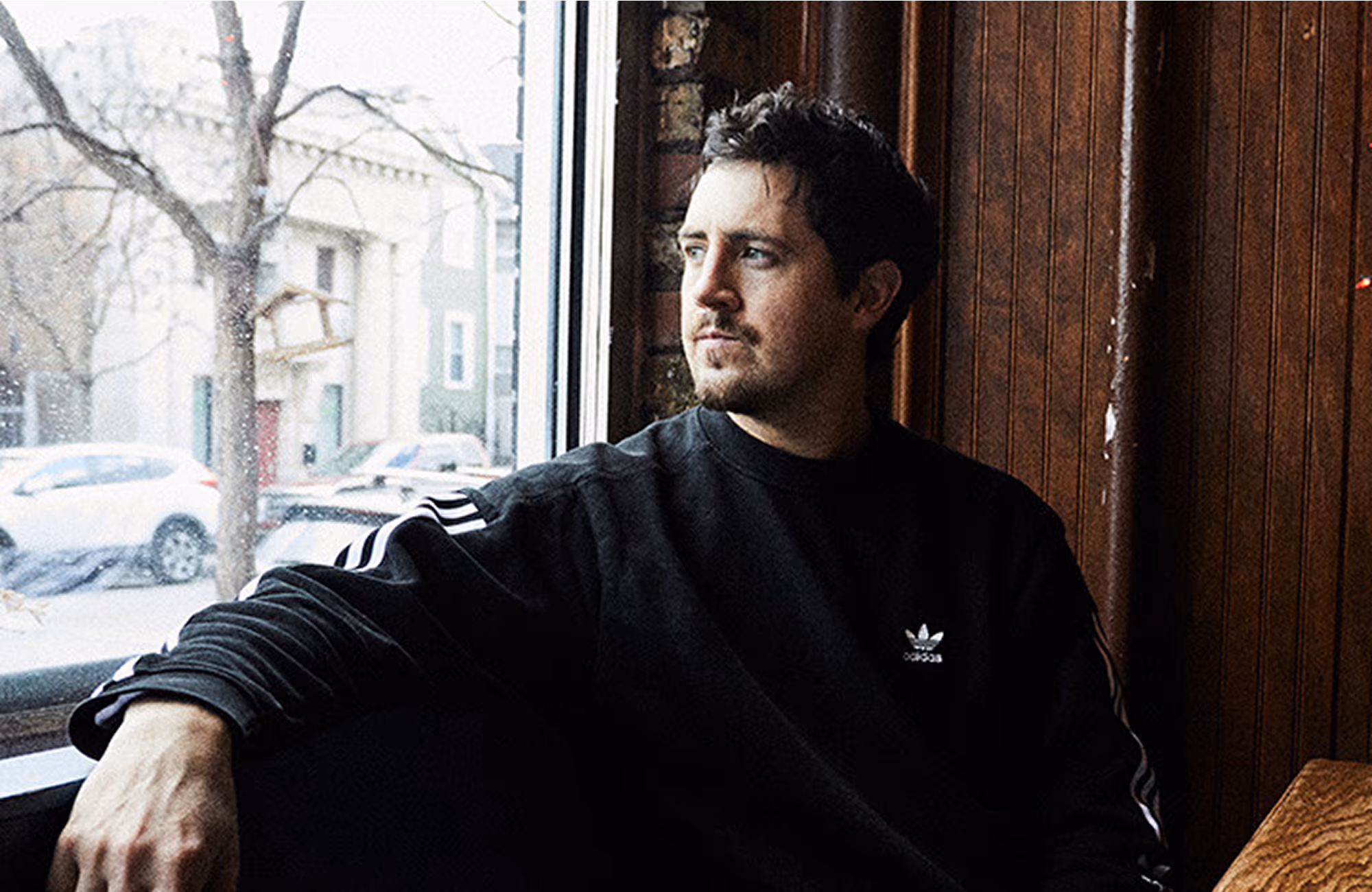 Man with dark hair and beard wearing a black Adidas sweatshirt, gazing out a window in a wooden-paneled room.