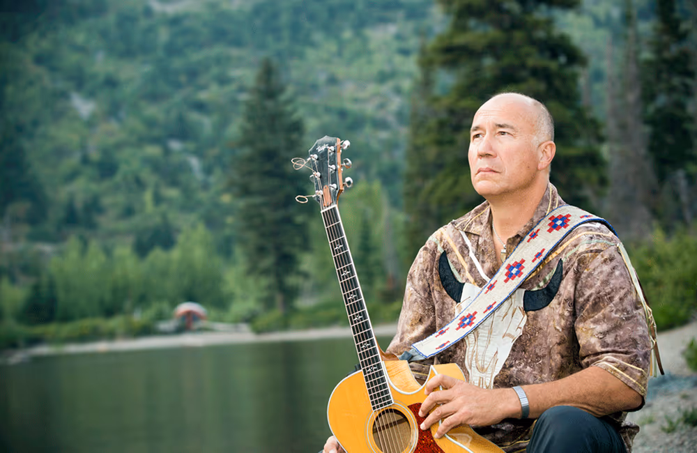 Man sitting outdoors with an acoustic guitar, wearing a patterned shirt and strap, with blurred trees and lake in the background.