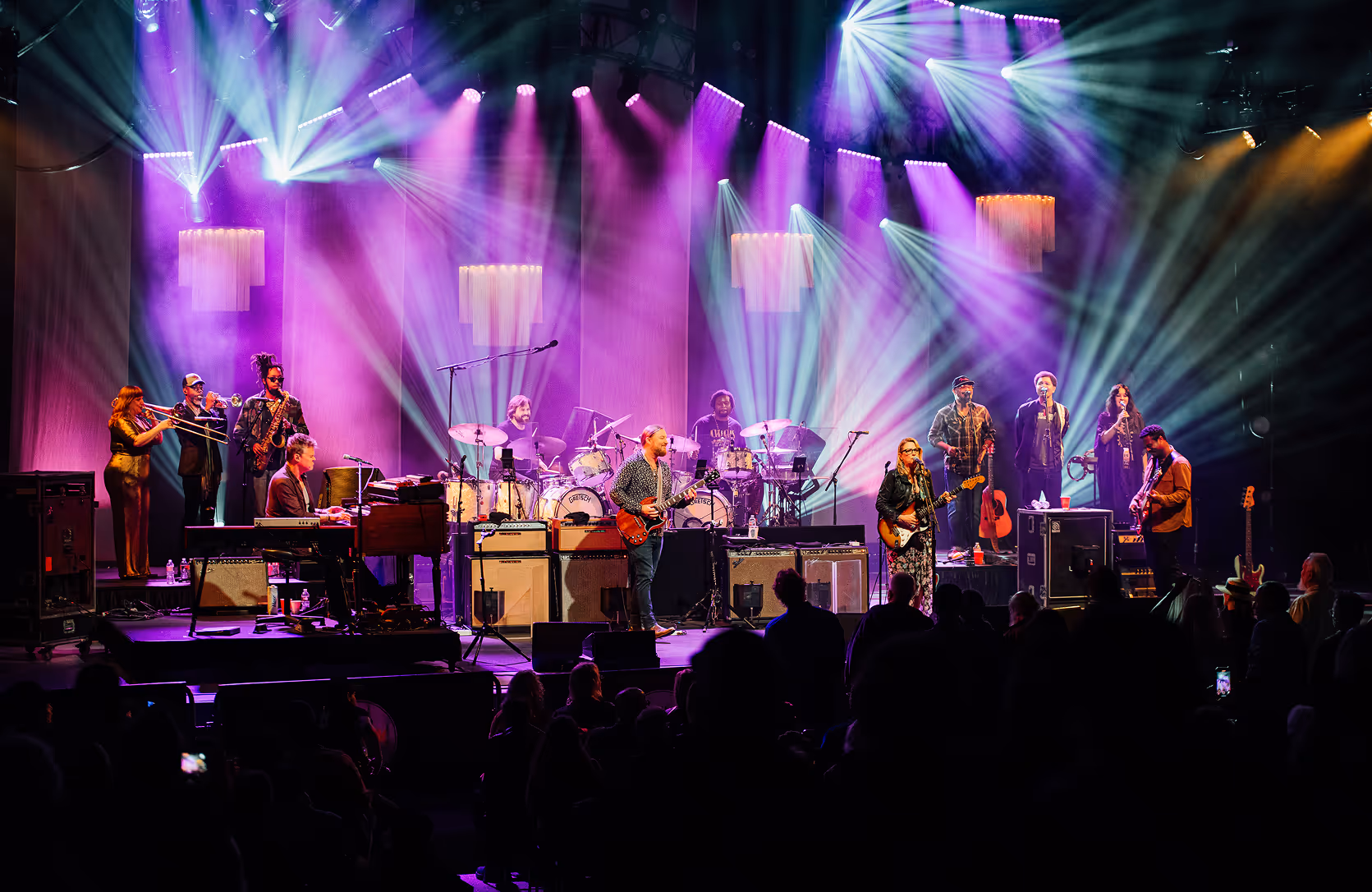 Band promo: Tedeshi Trucks band. Large band performing on stage with colorful purple and blue lighting and audience silhouettes in the foreground.
